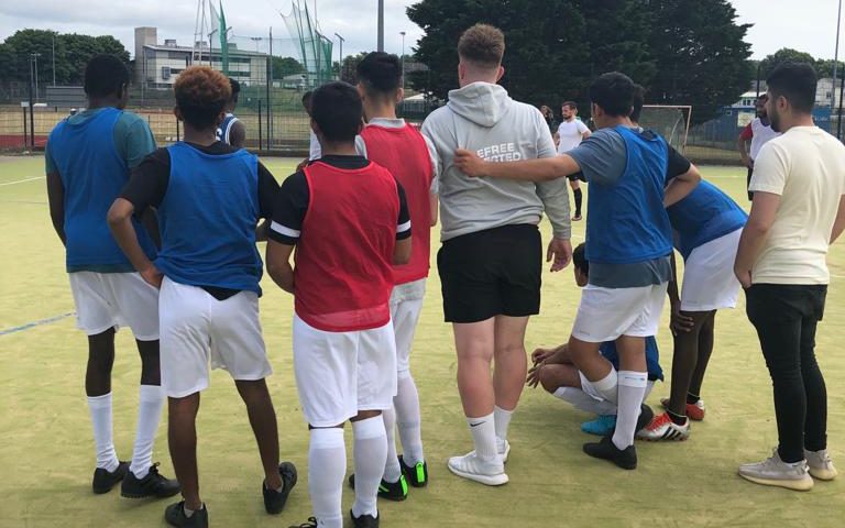 A group of 10 young men in football bibs gather round a youth worker in preparation for a football match.